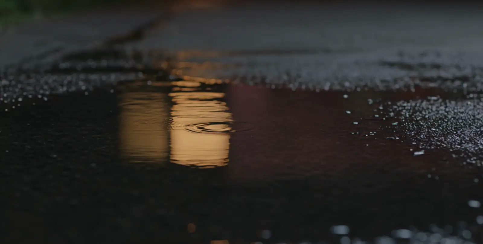 Rain falling into a shallow puddle on dark asphalt at dusk, a single amber streetlight reflected in the water